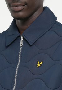 Marineblauer gesteppter Parka mit Reißverschluss, Punktkragen und einem gelben gestickten Logo auf der linken Brust. Glatte Textur mit Wellenmuster.