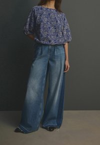 Modrá halenka s vzorem a nafouklými rukávy v kombinaci s džínovými kalhotami s širokými nohavicemi. Kalhoty mají roztřepený lem a různé odstíny modré.