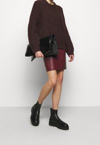 Weinroter Strickpullover kombiniert mit einem weinroten Leder-Minirock und schwarzen Stiefeletten. Das Modell hält eine glänzende schwarze Clutch.