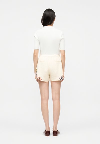 Femme debout de dos, portant un haut blanc à manches courtes, un short crème avec une broderie florale colorée, et des mocassins marron foncé.