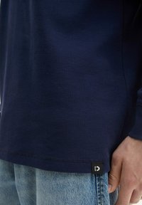 Marineblaue Langarmbluse aus strukturiertem Stoff mit einem subtilen Rautenmuster, versehen mit einem schwarzen Etikett mit einem weißen Logo am unteren Ende.