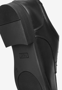 Chaussure en cuir noir avec un talon carré, semelle en caoutchouc texturée et coutures visibles. Le logo est embossé sur le côté. Fini lisse dans l'ensemble.