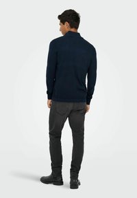 Pull en maille bleu marine avec un motif texturé, manches longues et col montant, associé à un jean gris foncé et des bottes noires.