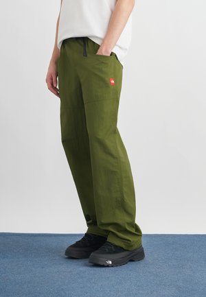 Persona con pantaloni verdi oliva con un logo rosso sulla tasca e scarpe da trekking nere che sta su un tappeto blu.