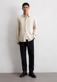 Helles beige Hemd mit Knöpfen, schwarze Hose und schwarze Schnürschuhe. Das Hemd hat eine leicht lockere Passform, während die Hose an den Beinen schmal zuläuft.