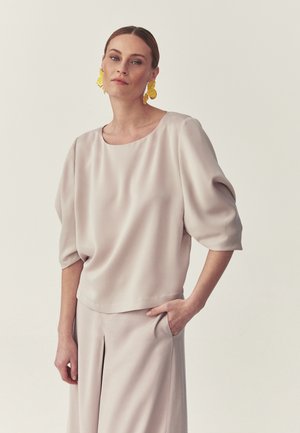 Kobieta ubrana w beżowy, luźny top z rękawami o długości do łokcia z bufkami oraz dopasowane, szerokie spodnie, uzupełnione dużymi żółtymi kolczykami w kształcie kwiatów.