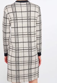 Robe grise avec un motif à carreaux noirs, manches longues et col côtelé noir. Le tissu semble doux avec une finition texturée.