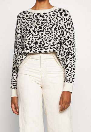 Mujer que lleva un suéter de estampado de leopardo en blanco y negro metido en pantalones de talle alto de color blanco roto, de pie frente a un fondo liso.