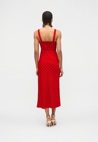 Γυναίκα με πλεξούδες, φοράει κόκκινο midi φόρεμα με μαύρες πουά και λευκά σανδάλια με ψηλό τακούνι, στέκεται με την πλάτη στο λευκό φόντο.