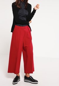 Pantalons larges rouges avec des plis, associés à une chemise noire à boutons et un cardigan noir. Des chaussures noires avec un accent de semelle blanche.
