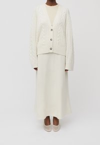 Cárdigan de punto trenzado en crema con escote en V y tres botones, superpuesto a una falda larga de crema a juego y zapatos planos. Tejido suave y texturado.
