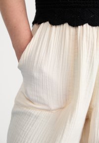 Light cream pants with textured, pleated fabric. Features side pockets, a relaxed fit, and a black crocheted waistband with scalloped edge.