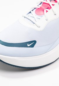 Detailní záběr na bílé a světle modré Nike tenisky s růžovým akcentem a viditelnými tkaničkami na bílém pozadí.