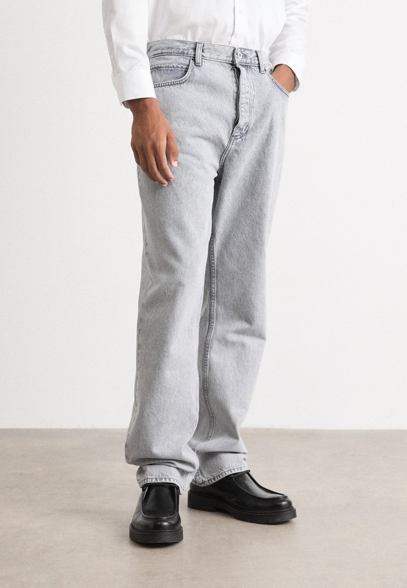 Lys grå denimjeans med rett ben-design, subtil tekstur og knappestang-lukking, kombinert med svarte slip-on sko.