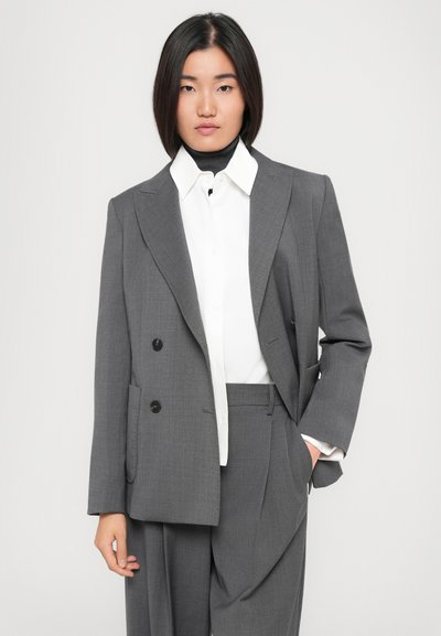 Blazer grigio doppiopetto con rever a punta, bottoni neri, tessuto textured, indossato sopra una camicia bianca a collo, abbinato a pantaloni grigi coordinati.