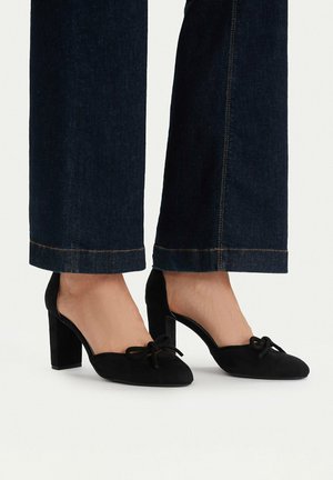 Füße in schwarzen Wildleder-Pumps mit Blockabsatz und Schleifendetail, kombiniert mit dunkelblauen Jeans, die über dem Knöchel gekürzt sind.