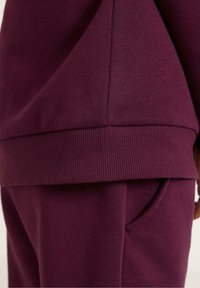 Burgunderfarbener Sweatshirt mit geripptem Saum und Bündchen, präsentiert mit passenden Hosen, die eine Seitentasche und eine glatte Stoffstruktur aufweisen.