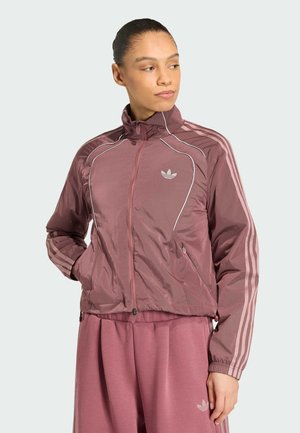 Frau trägt einen mauvefarbenen Adidas Trainingsanzug mit Reißverschlussjacke und passenden Hosen, posiert mit den Händen in den Taschen und schaut zur Seite.