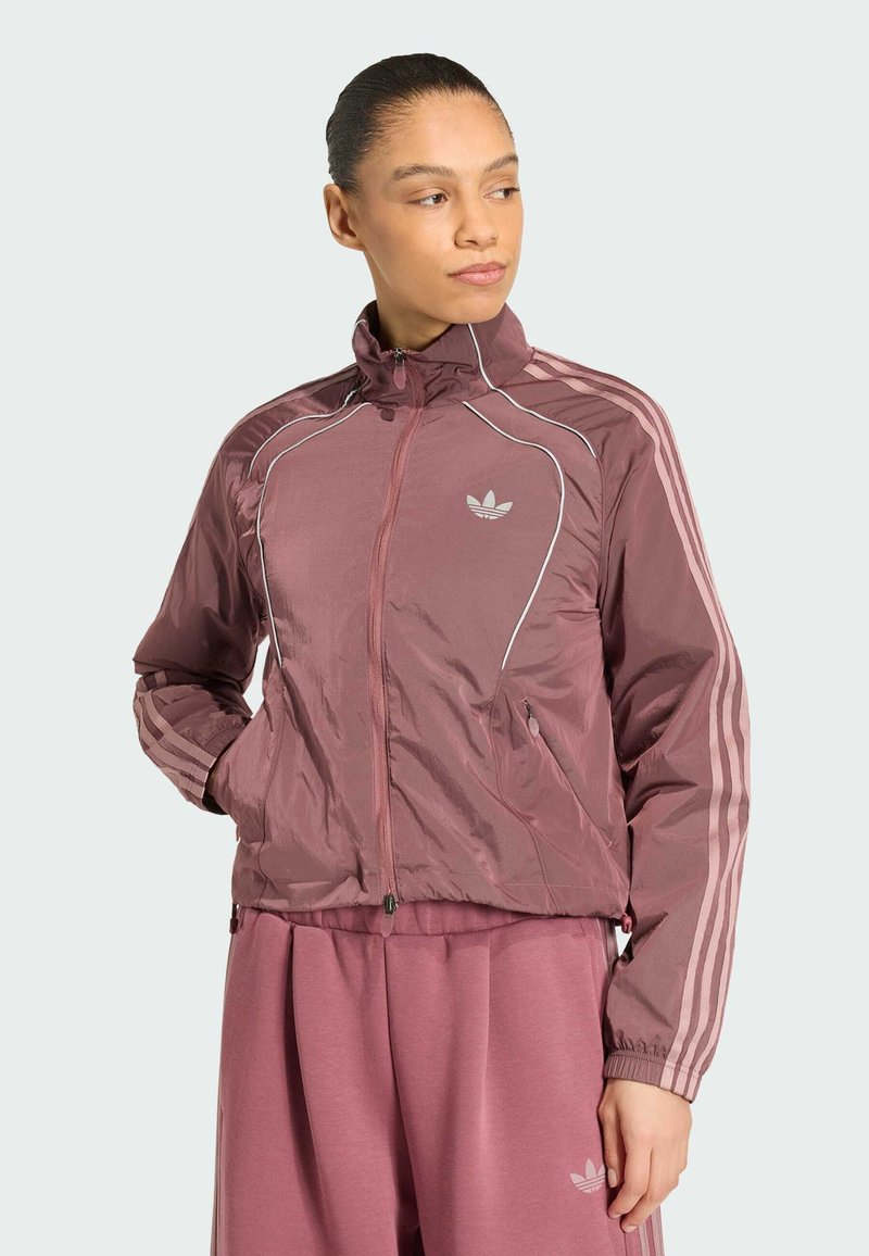 Femme portant un survêtement mauve Adidas avec une veste zippée et un pantalon assorti, posant avec les mains dans les poches, regardant sur le côté.
