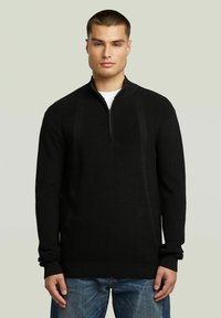 Un pull à côtes noir avec un col zippé mi-hauteur, des manches longues et un motif texturé. Associé à un jean en denim bleu.