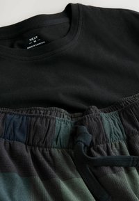 Camiseta de algodón negra parcialmente colocada sobre pantalones de salón con estampado en tonos oscuros de verde y negro, que cuentan con una cinturilla elástica y cordón.