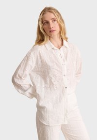 Blusa de algodón blanca con diseño de ojales que presenta patrones cuadrados, con botones en el frente y un cuello clásico. Mangas largas y corte relajado.