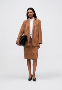 Žena u smešnom antilop blejzeru smeđe boje i suknji s belom košuljom, drži veliki crni clutch dok stoji na svetlosivoj pozadini.
