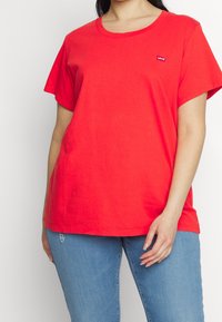 Femme portant un t-shirt rouge à manches courtes Levi's et un jean déchiré bleu clair, se tenant devant un fond blanc uni.