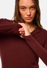 Jeune femme ajustant la manche de son pull à manches longues bordeaux, regardant directement l'objectif.