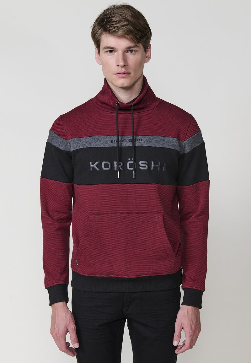Sudadera roja y negra con un alto cuello, que presenta una tela texturizada, bolsillo frontal y el logo de KORÖSHI en el pecho con letras en negrita.