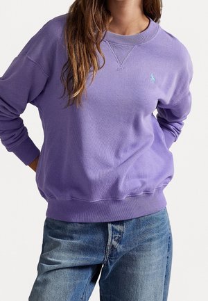 Kobieta ubrana w jasnofioletową bluzę z okrągłym dekoltem z małym haftowanym logo, w połączeniu z niebieskimi dżinsami, stoi z rękami za plecami.