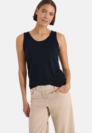 Femme portant un haut noir sans manches et un pantalon beige, la main gauche dans la poche, debout devant un fond blanc uni.