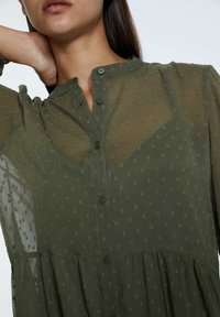 Durchsichtige olivgrüne Bluse mit strukturierten Punkten, Knopfverschluss vorne, rüschigem Kragen und lockerer Passform, mit einem gestuften, fließenden Design.