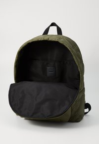 Sac à dos matelassé vert avec un intérieur noir. Présente un sommet arrondi, un tissu robuste et une petite étiquette intérieure. Design ouvert pour un accès facile.