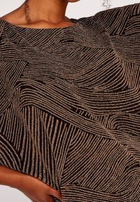 Haut en tissu noir avec des lignes ondulées dorées et bronze. Texture douce. Coupe carrée avec un col bateau et des manches larges.