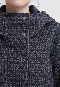 Marinblå jacka med geometrisk design, mjuk textur och huva. Fem silverknappar fram ger detaljer.