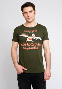 Camiseta verde oliva con un gráfico de Wile E. Coyote, texto "Warner Bros. Presents" y jeans negros desgastados. Mangas cortas y cuello redondo.