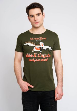 Olivově zelené tričko s grafikou Wile E. Coyote, textem "Warner Bros. Presents" a poničenými černými džíny. Krátké rukávy a kulatý výstřih.