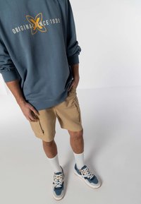 Modrá mikina s potlačou "ORIGINAL SINCE 1939", béžové cargo šortky s bočnými zipsami, biele ponožky a modré tenisky so šedými akcentmi.