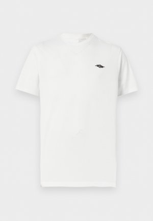 Jeune homme aux cheveux bouclés portant un t-shirt blanc uni à manches courtes avec un petit logo noir sur la poitrine et un pantalon cargo noir.