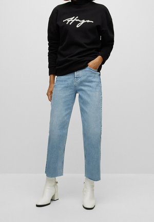 Persoon draagt een zwarte Hugo-trui, lichtblauwe cropped jeans met rafelige zomen en witte enkellaarsjes met blokhakken.