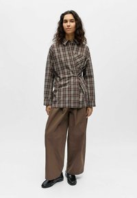 Chemise à carreaux dans des tons terreux, avec un design asymétrique et un lien sur le côté. Associée à un pantalon ample marron et des chaussures noires.