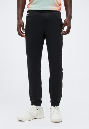 Mann trägt schwarze Sporthose und weiße Sneakers, steht vor einfarbig hellem Hintergrund.