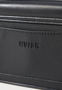 Svart faux läderhandväska med texturerad yta, pryds av det präglande "HVISK"-logotypen och fin sömnad längs kanterna.