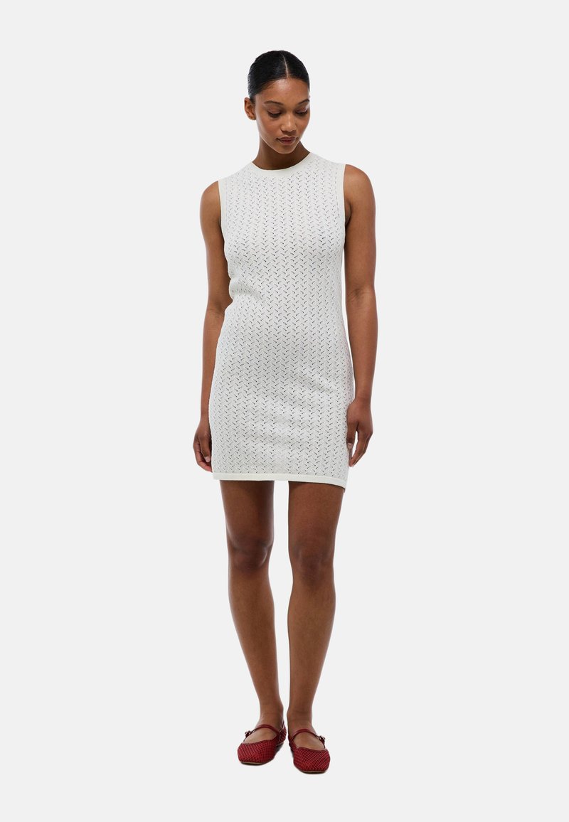 Robe blanche sans manches avec un motif de tricot texturé, courte, et un col rond. Associée à des chaussures plates rouges.