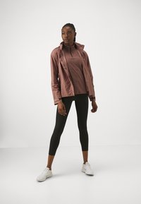 Giacca a vento leggera di rosa polveroso sopra un top a zip quarti marrone scuro, abbinata a leggings neri e scarpe da ginnastica bianche.