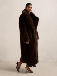 Brun faux fur-kåpe med en bred, polstret krage, lange ermer og kne-lengde design. Bært over en mørk kjole og kombinert med svarte hælsandaler.