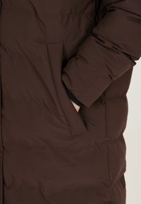 Brun puffer-jakke med en tekstureret overflade, der har en sidelomme og elastiske manchetter. Stoffet ser tykkere og isoleret ud.