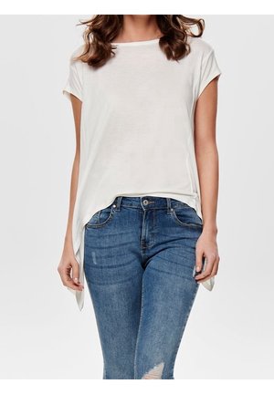 Femme portant un haut blanc ample à manches courtes associé à un jean skinny bleu déchiré, debout devant un fond uni.