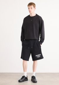 Black sweatshirt with a logo patch, paired with black shorts featuring white "HUGO" text. Model wears black sneakers and white socks.